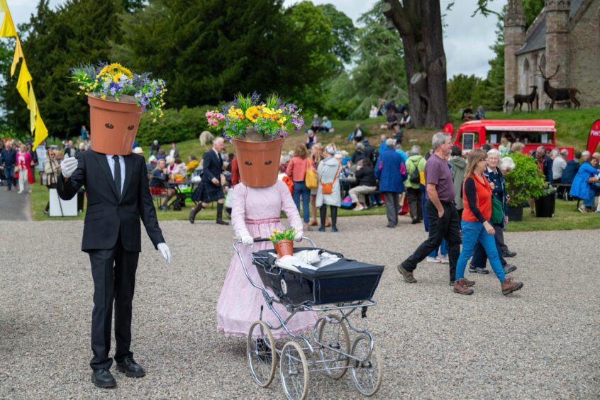 Suited man with flower pot for head beside woman with flower pot for head pushing pram