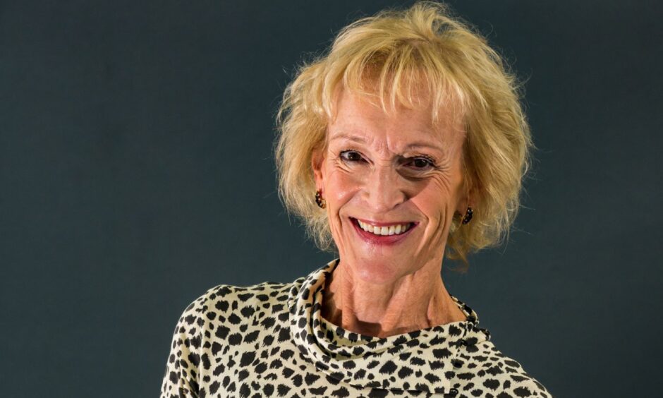 Image shows: author and cookery writer Sue Lawrence. Sue is wearing a black and white spotted dress and is standing with her arms loosley folded around her waist and is smiling warmly at the camera. She has blonde hair cut in a short bob with a fringe.