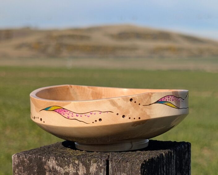 Image shows: a hand-turned wooden bowl designed and made by Debbie Coull, Arncroach. The bowl is sitting on a fence post with fields and a hill in the background.