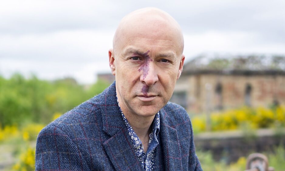image shows author Chris Brookmyre who is taking part in the Dundee Book Festival. He is standing in the gardens of a large house and is wearing a dark blue jacket and patterened shirt. He is bald and has a port-wine birthmark on his nose.