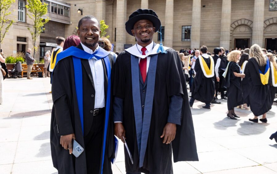 Two male graduates pose in the sunshine. 