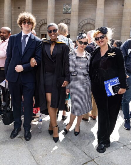 A female graduate flanked on either side by proud friends and family members, all wearing sunglasses. 