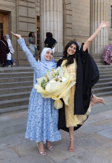 Two female graduates throw their hands in the air outside Caird Hall. 