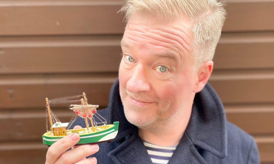 Image shows: author and broadcaster Charlie Connelly is holding a tiny wooden model boat. He is wearing a striped Breton-style t-shirt and navy jacket. Charlie is taking part in the Dundee Book Festival 2025.