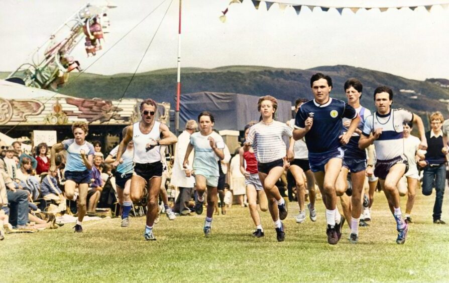 A group of runners approaching the finish line, with bunting and rides in the background and a hill in the distance.