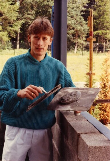 Baltacha holding a trowel and a board with cement on while constructing a brick wall at his home.