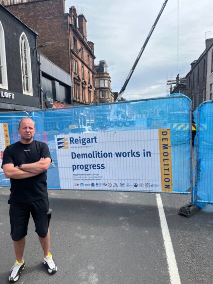 Craig Fraser, arms folded, beside metal barrier with sign saying 'demolition works in progress'