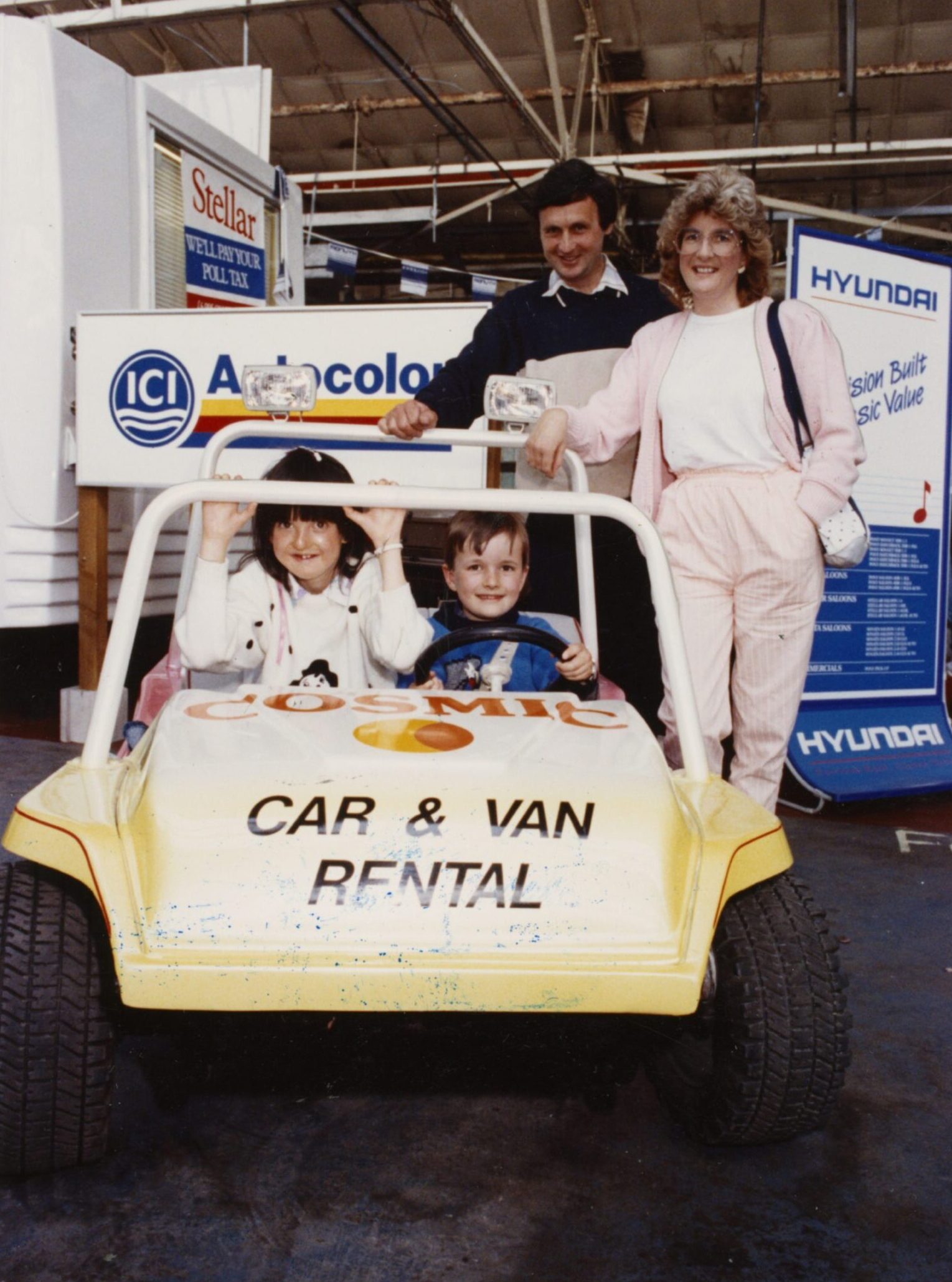 two kids, watched by their parents, sit inside a pedal car