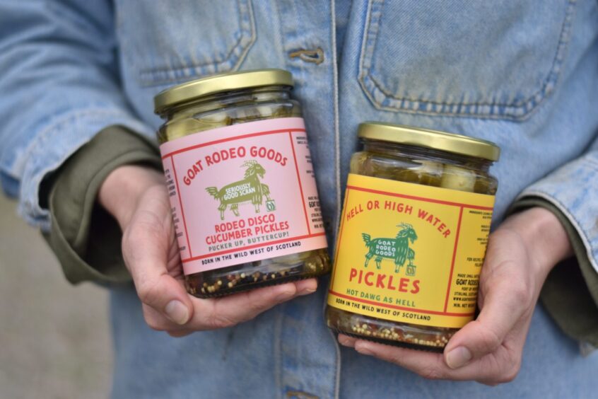 A close-up of a person holding two jars of Goat Rodeo Goods pickles against their denim jacket.
