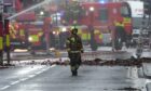 Firefighter crossing street with rubble, fire crews and appliances behind