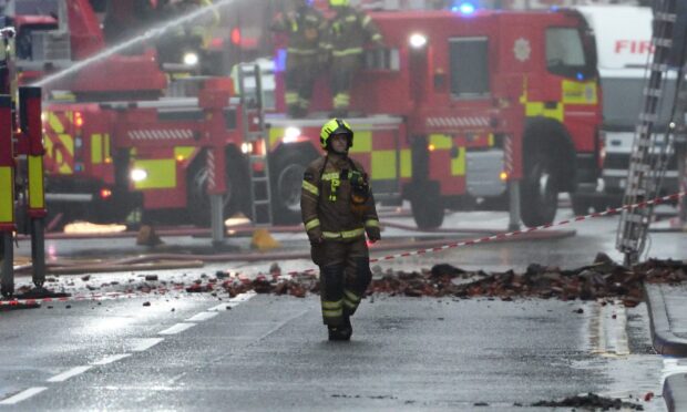 Firefighter crossing street with rubble, fire crews and appliances behind