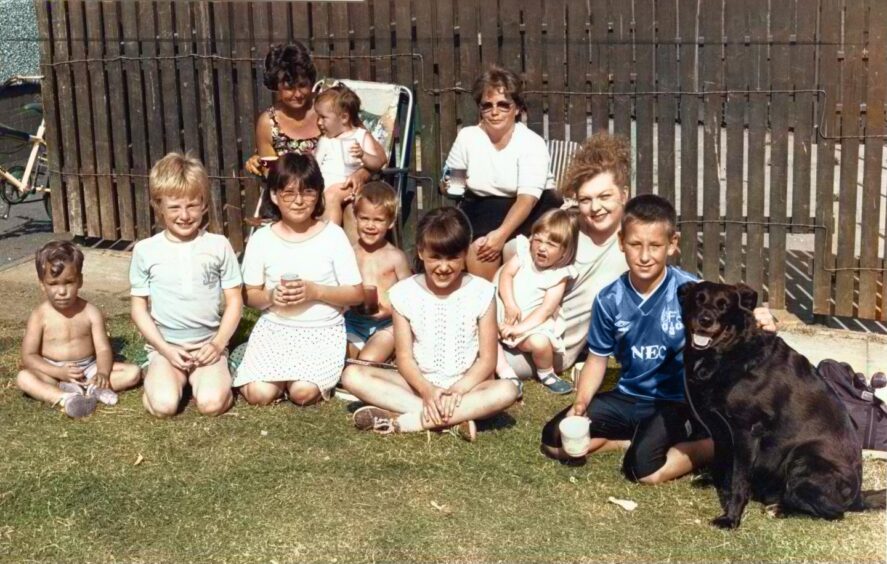 Two women sit on deckchairs in the sun, joined by several children and a black dog.