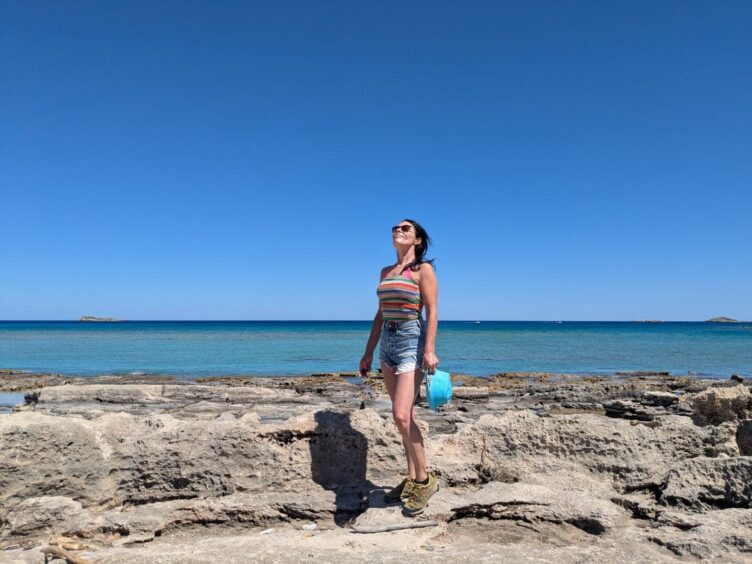 Gayle standing on a rocky shore in Rhodes with the sea behind her. 