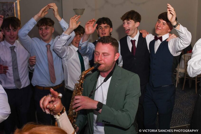 A man playing a woodwind instrument to the encouragement of departing pupils. 