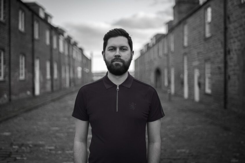Image shows: singer/songwriter Jamie Button. He is standing in the middle of a street with terraced houses disappearing into the distance. He is wearing a black t-shirt and has black hair and beard and a serious expression.