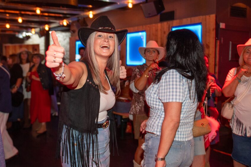 A woman in a cowboy hat and tasselled waistcoat gives the thumbs up at a nightclub. 