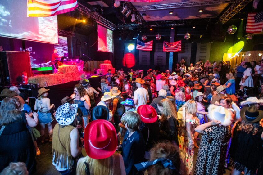 Revellers, many wearing cowboy hats, in a nightclub whose walls are hung with USA flags. 