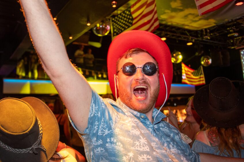 A man in shades and a red cowboy hat in a nightclub. 