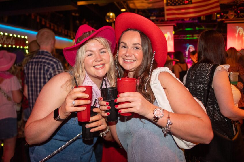 Two women dressed in denim and red cowboy hats clutch drinks in a nightclub. 