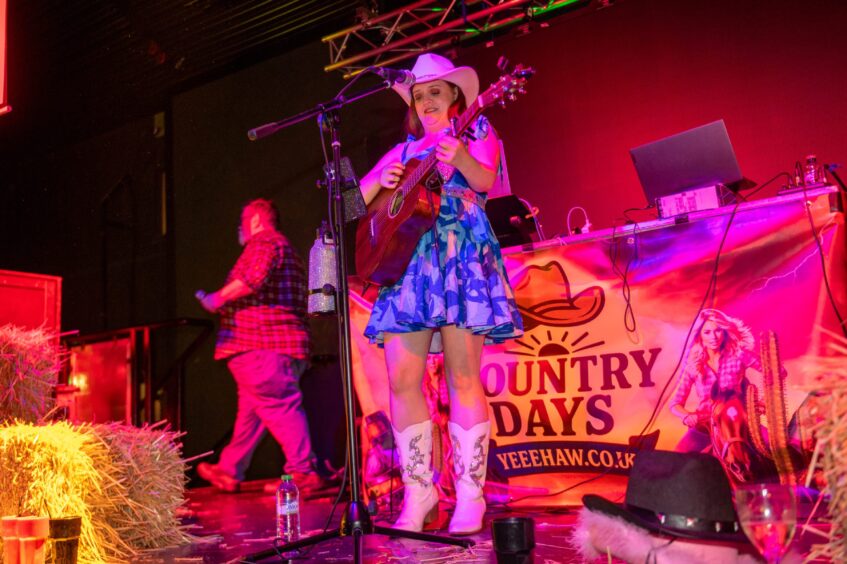 A woman dressed in country and western garb plays a guitar on a nightclub stage. 