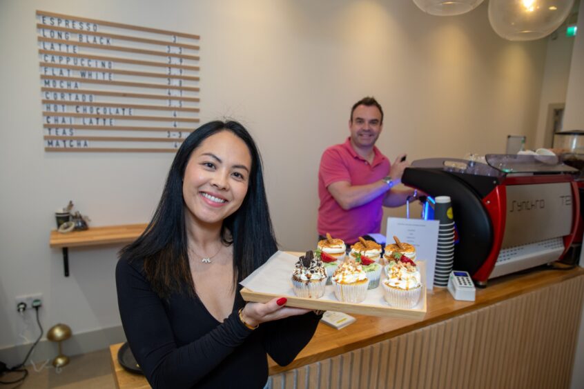 Lisa holding a tray of cakes while Lindsay makes coffee