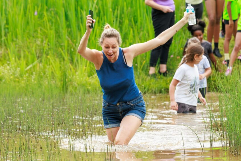 A woman holds her phone and water aloft as she wades through a muddy pond. 