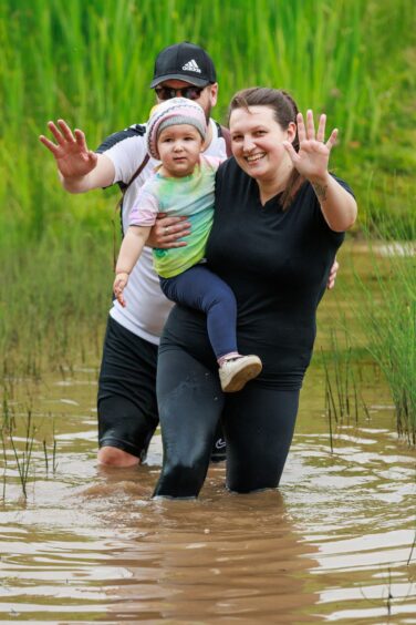 A man and a woman holding a child wave as they wade through a muddy puddle. 