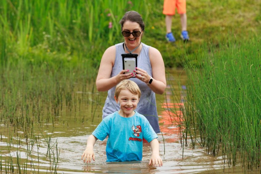 A woman takes a photo of a boy in front of her, both in a muddy pond. 