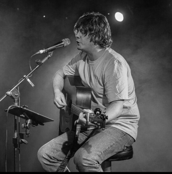 Image shows: singer/songwriter Pete Smith who is taking part in Dundee Music Festival. The black and white photo shows Pete on stage sitting on a stool with an acoustic guitar. He's wearing jeans and a t-shirt.