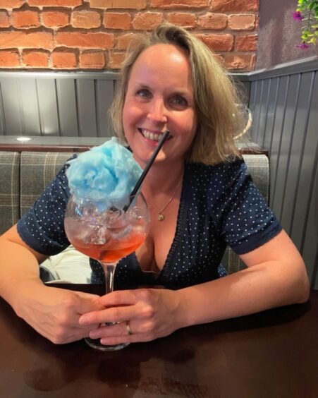 Journalist Rachel smiles at the camera while holding the stem of a large glass with a red cocktail in it and a large pile of blue candyfloss on the top.