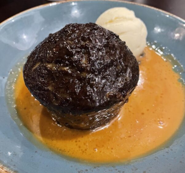 A plate with a large round portion of sticky toffee pudding surrounded by sticky toffee sauce and with a small scoop of vanilla ice cream.