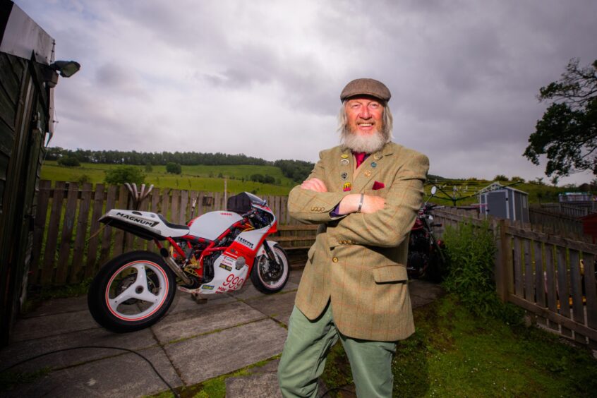 Geoff Mumford in tweed jacket, standing arms folded in front of motorbike