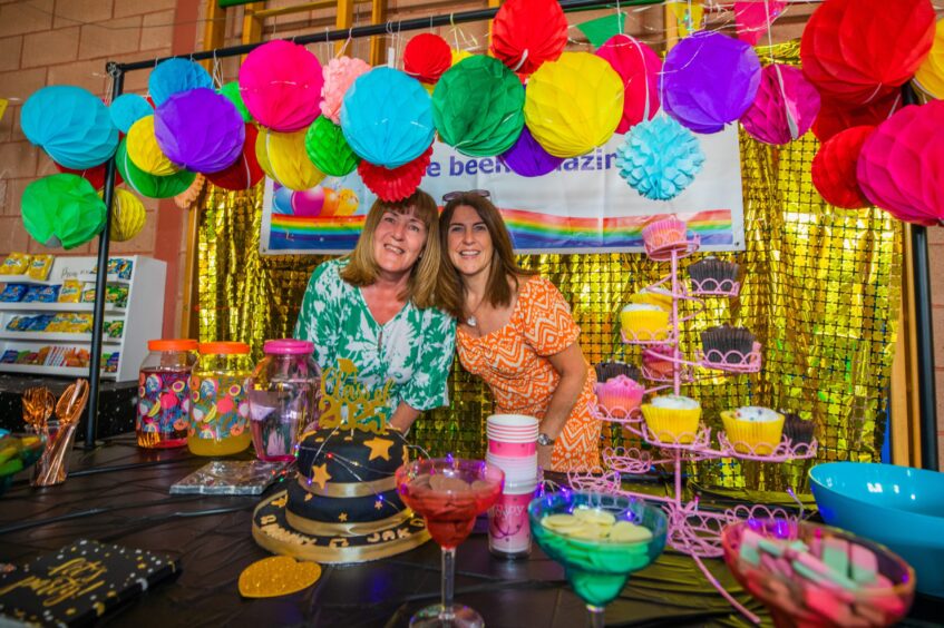 Two women at a vibrantly decorated stall containing cakes and juice. 