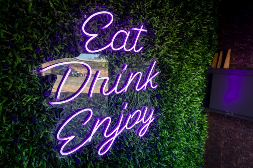 A purple neon sign saying Eat, Drink, Enjoy against a green background of fake foliage.