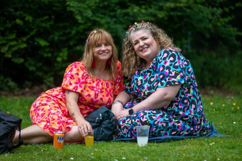 Karen Lessles and Jo Hastings smile and pose with flower garlands in their hair. They are sitting on a picnic blanket.