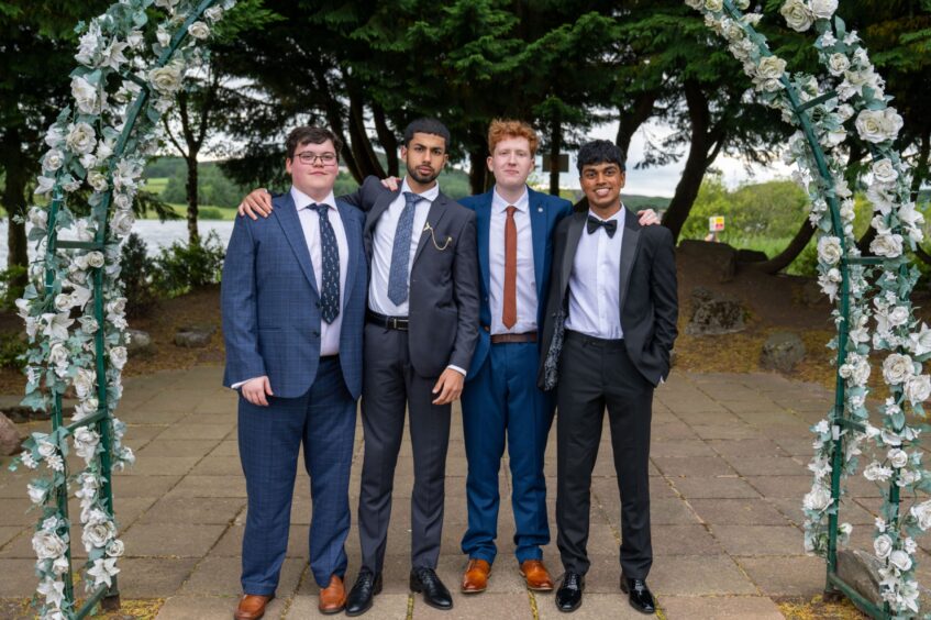 Four boys in suits standing under an arch decorated with flowers. 