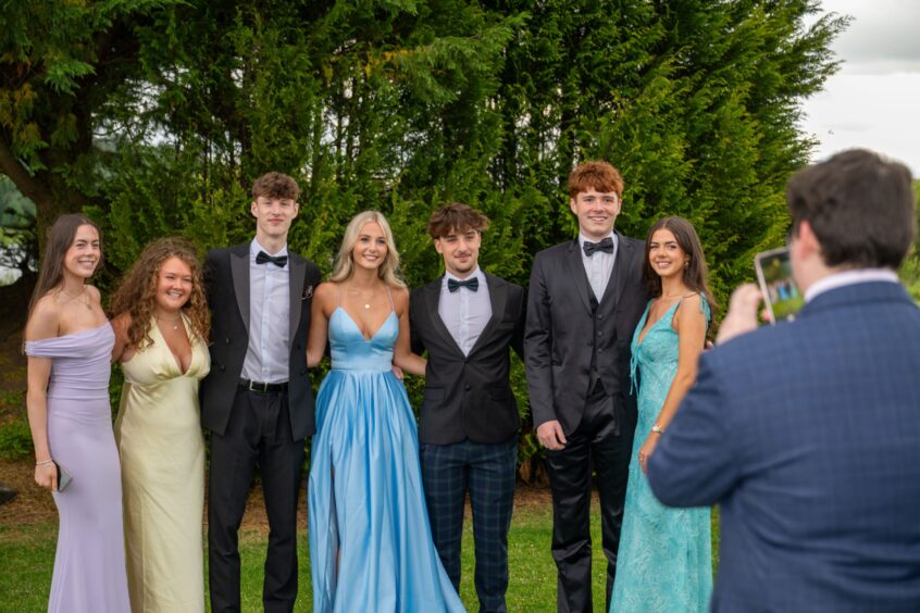A group of boys in dinner suits and girls in dresses outside. 