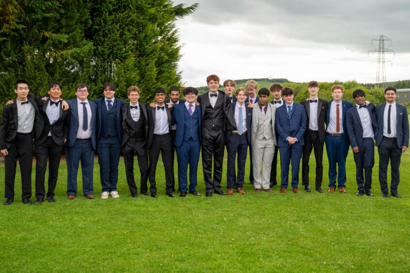 A line of boys in suits standing on a lawn with trees behind them. 