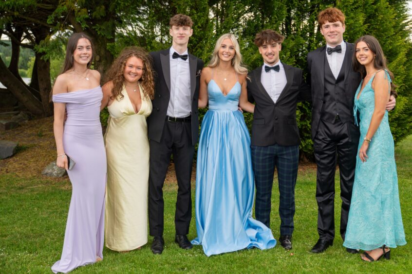A group of boys in dinner suits and girls in dresses outside. 