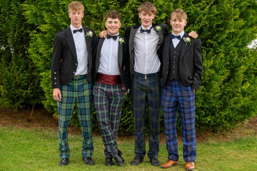 Four boys in white shirts, black suit jackets, bow ties and tartan trousers posing outside. 