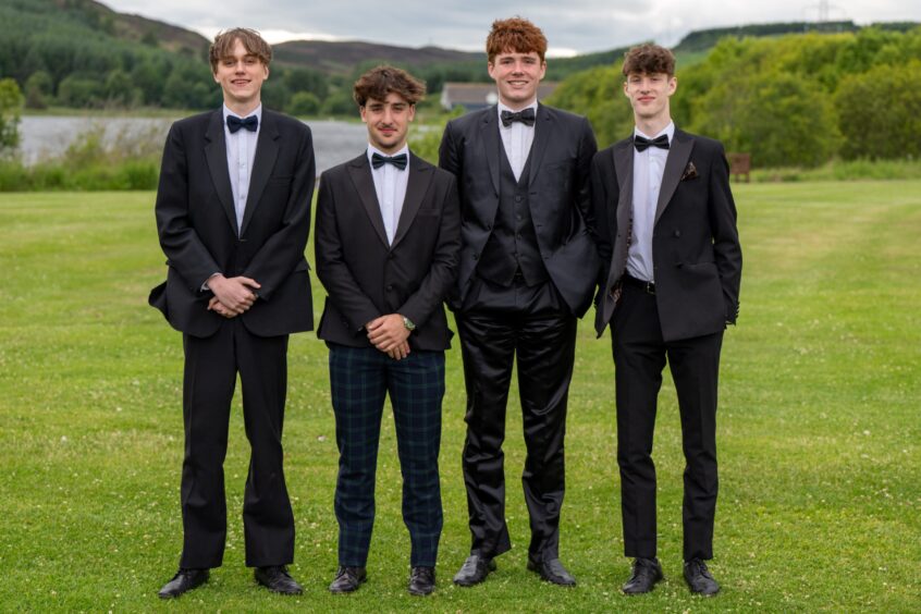 Four boys in dinner suits standing on a lawn with Piperdam Loch behind them. 