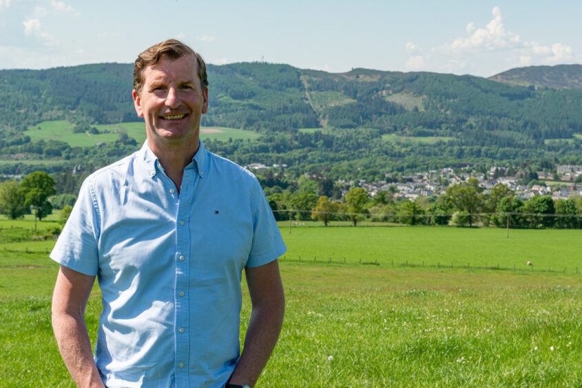 Dave Doogan in short sleeved pale blue shirt, standing in field with Perthshire landscape behind