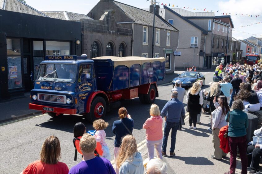 Classic blue and red Watson Bros vintage truck drives in Carnoustie gala day parade, watched by spectators lining the High Street on a sunny Saturday.
