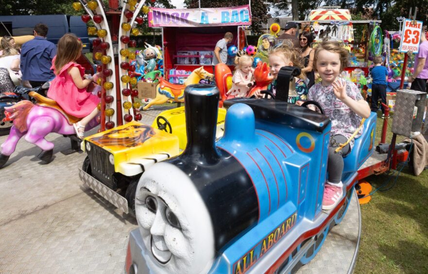 Daisy Dorrington and Darcy McGoldrick, both aged 3, wave and smile as they ride a blue train at the 2025 Carnoustie gala day fairground.