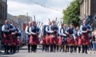 Pipe band dressed in kilts and blue shirts marches down Carnoustie High Street during the 2025 gala day parade as spectators watch and cheer.