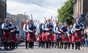 Pipe band dressed in kilts and blue shirts marches down Carnoustie High Street during the 2025 gala day parade as spectators watch and cheer.