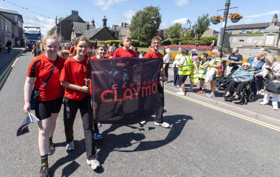 Carnoustie Claymores group in matching red shirts walk with their banner down the High Street during the 2025 gala day parade, watched by a large crowd.