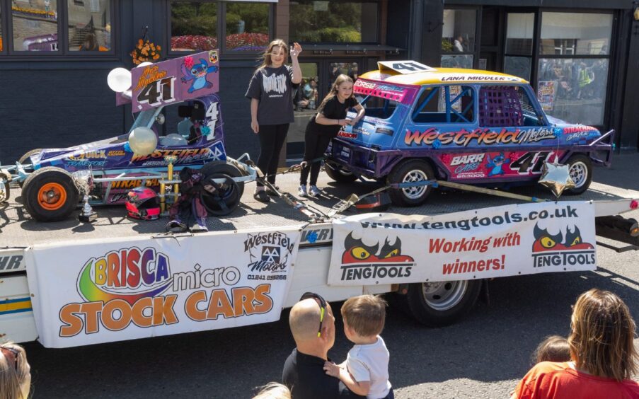 Two girls stand beside decorated BRISCA micro stock cars on a float sponsored by Westfield Motors during Carnoustie gala day 2025.