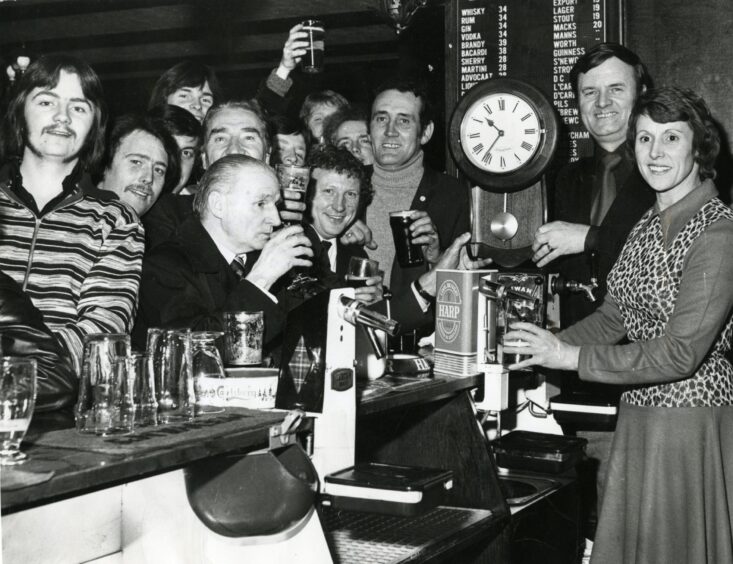 tow people pour drinks behind the bar as patrons lean in for a picture