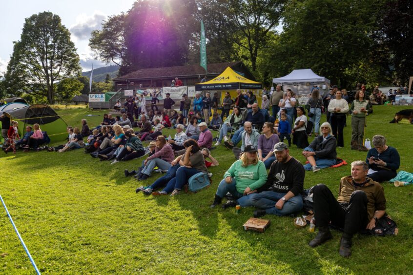 Crowd members seated and standing on grass watching games with tents behind